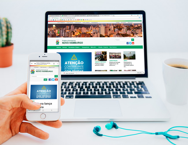 Ao fundo, a imagem de um notebook e, à frente, uma mão segura um celular com a imgem do novo site