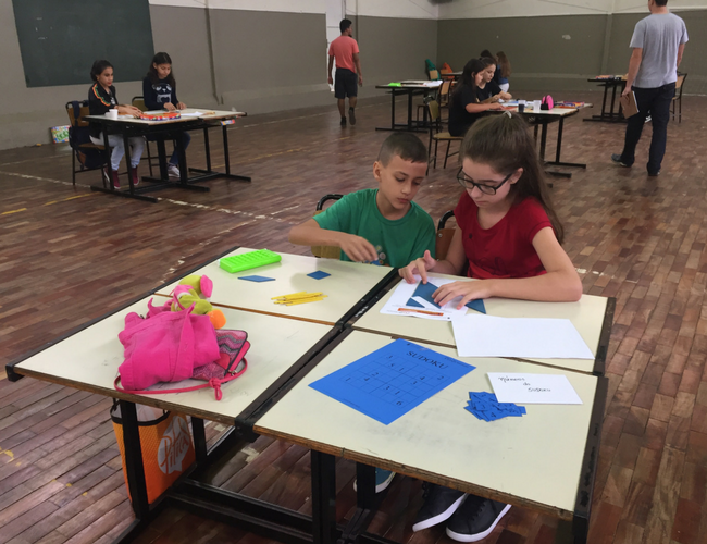 Duas crianças sentadas desenvolvem uma atividade com papíes em cima de classes escolarres