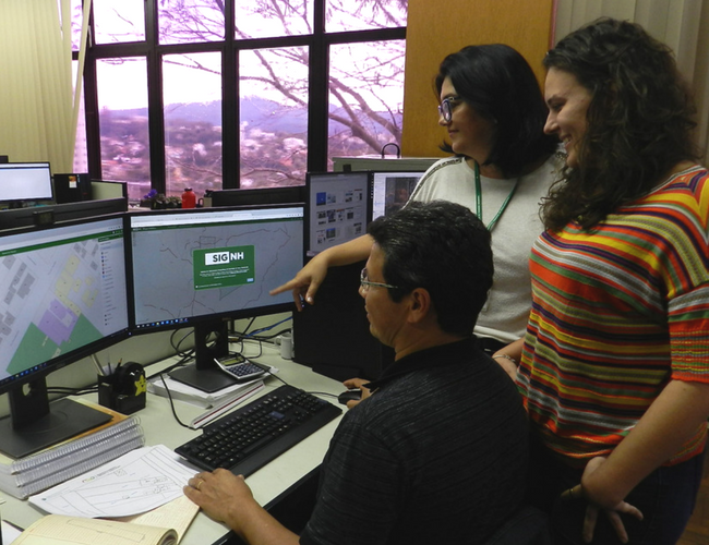 Um homem sentado e duas mulheres em pé olhando para um monitor de computador