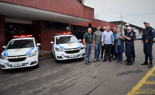 Dois carros parados à esquerda e a prefeita e equipe em pé à direita