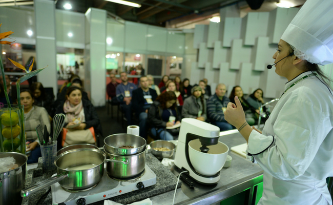 Uma mulher em pé de branco cozinha enquanto o público sentado assiste