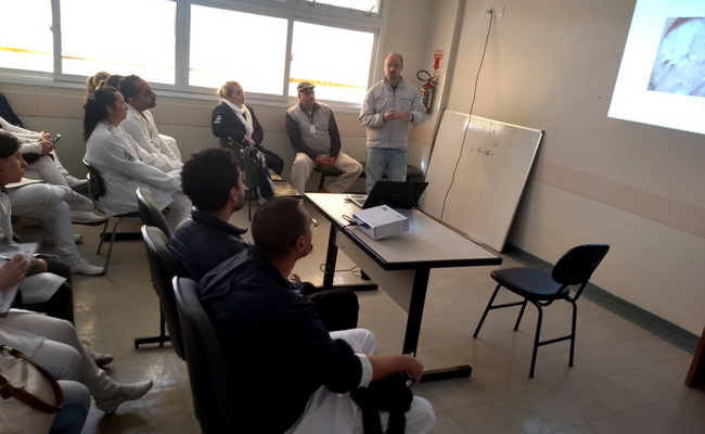 Um homem em em pé (professor) e os alunos sentados em uma sala de aula