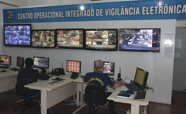 Dois homens sentando monitoram cinco televisões com imagens captadas pelas câmeras nas ruas