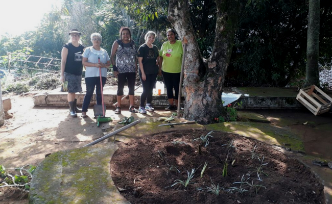 Cinco mulheres que formam o grupo em pé em frente a uma das floreiras plantadas