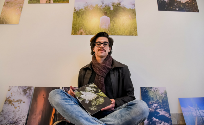 O artista Tom Ferreiro sentado com as obras ao fundo