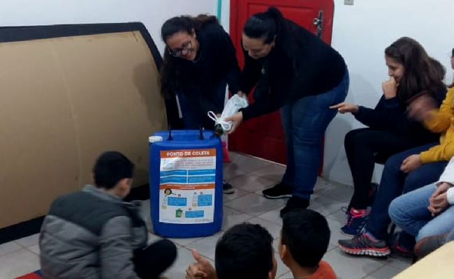 Com um tonel azul ao centro, duas professora em pé e alunos sentando ao entorno do tonel  