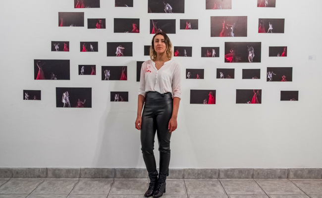 Artista France em pé com os quadros expostos atrás em uma parede branca
