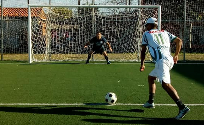 Ao fundo, o goleiro parada no meio da goleira e um jogador na marca do penalti correndo ao encontro da bola