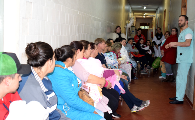 À direita, o pediatra em pé. À esquerda, as mamães e bebês sentados