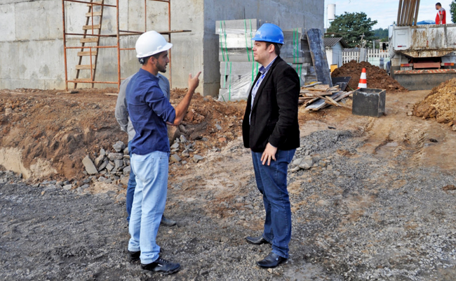 Dois homens em pé vistoriando o canteiro de obras, formado por um prédio de concreto