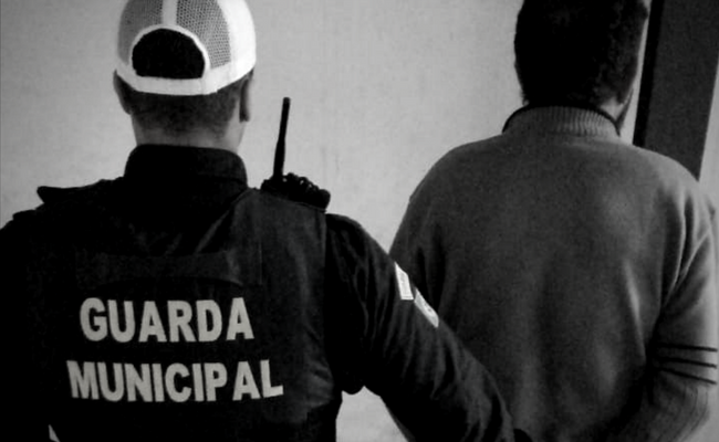 Dois homens de costas. À esquerda, o Guarda Municipal e, à direita, o preso