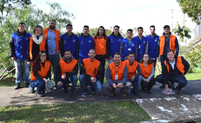 Grupo de pessoas identificadas com coletes laranjas e azuis. Um grupo está em pé e outro agachado