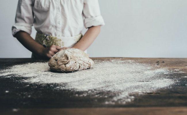 Em uma mesa preta, um padeiro amassa uma massa de pão repleta de farinha