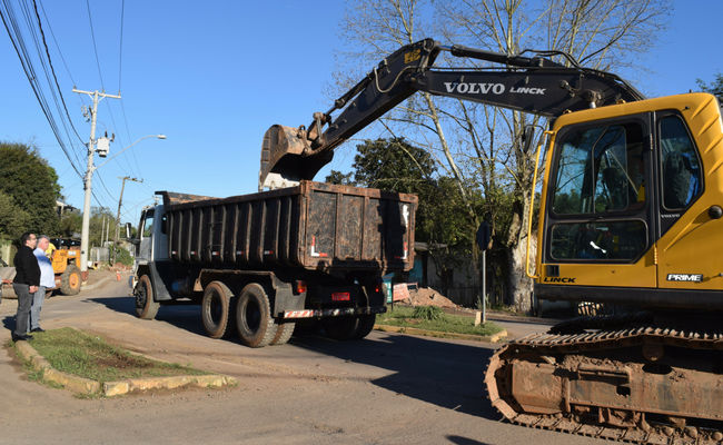 Uma caminhão parado à esquerda com uma retroescavadeira à direita largando o material na caçamba do caminhão