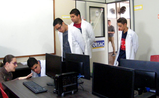 Em uma sala com computadores, quatro alunos em pé e de jalecos brancos, vistoriam a sala
