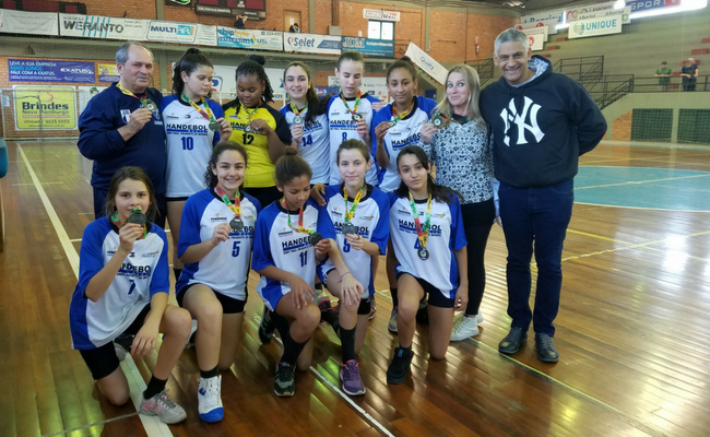 Em uma quadra de handebol, três menina e o treinado em pé e quatro meninas sentadas 