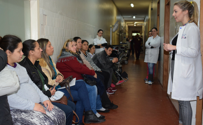 Em um corredor da maternidade, em pé, à direita, psicóloga fala para as gestantes, que estão sentadas à sua frente 