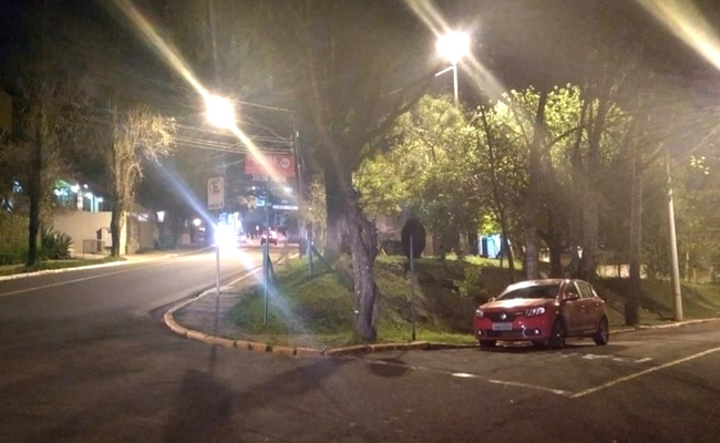 Duas ruas. No meio a praça com grama verde e dois postes com lâmpadas acessas