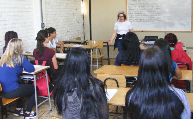 No fundo, a professora em pé. Sentadas seis alunas em sala de aula