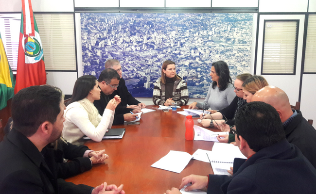Em uma mesa, com a prefeita sentada na ponta, depois pessoas também sentadas no entorno