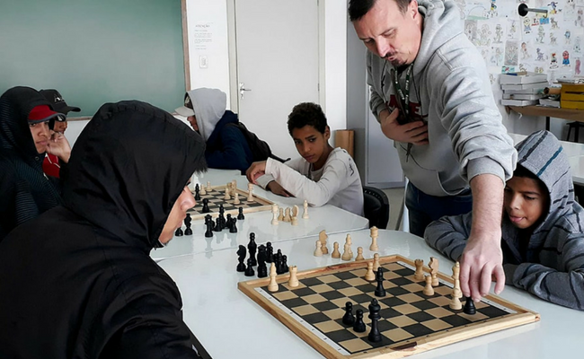 Em uma mesa, ao centro, uma tabuleiro de xadrez. De cada lado, um jogador sentado. Em pé, o professor orienta os alunos mexendo no tabuleiro