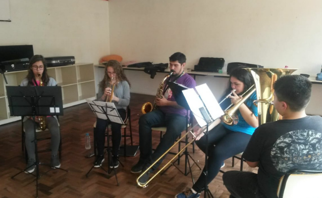Em um semicírculo, crianças estão sentadas tocando instrumentos