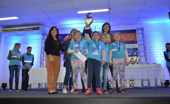 Em um palco, três crianças seguram um troféu ao lado da secretária Maristela Guasselli