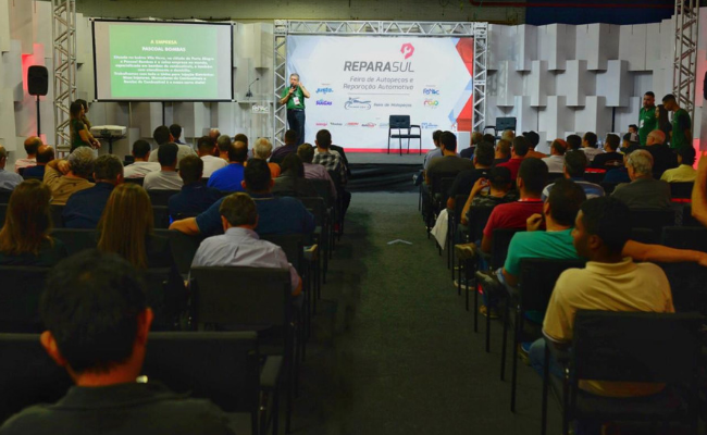 Ao fundo, um palco com um palestrante e o público sentados em cadeiras