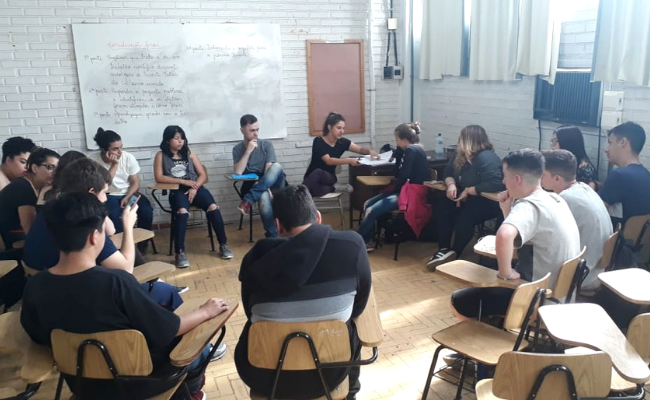 Em uma sala de aula, estudantes estão sentados em cadeiras em círculo