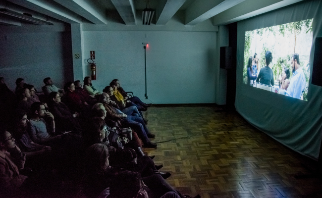À esquerda, público sentando em cadeiras. À direita, em um pano branco, o filme projetado