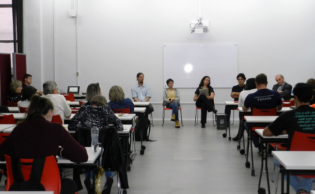 Em uma sala, ao fundo, três pessoas sentadas em cadeiras. Em frente a estas pessoas, o público sentado lado a lado também em cadeiras