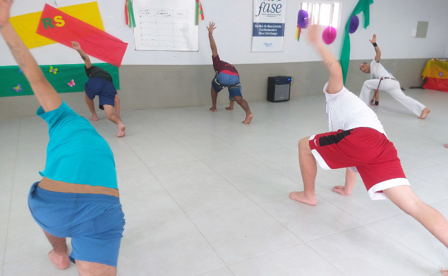 Em uma sala, cinco homens de costas fazem um movimento de capoeira com uma perna e um braço escorados no chão