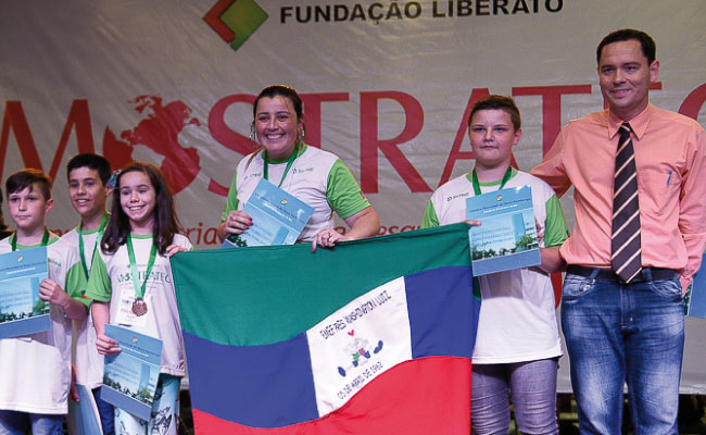 Em pé em um palco, alunos seguram a bandeira da escola e os prêmios