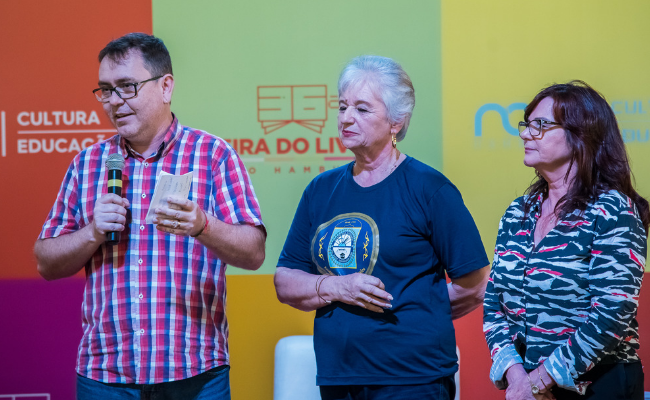 Secretário Ralfe em pé ao microfone. Ao lado, também em pé, Renate Gigel e a secretária Maristela