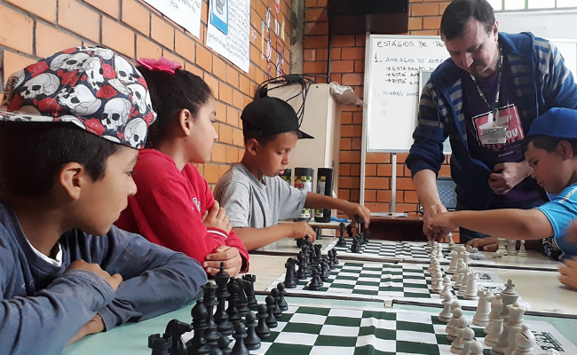 Em uma mesa, à esquerda, três meninos sentandos. Sob a mesa, três tabuleiros de xadrez com as peças. Ao fundo, em pé, o instrutor de xadrez