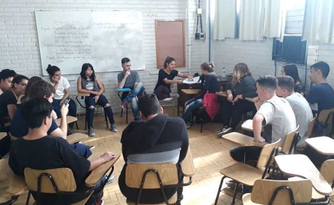 Em uma sala, alunos sentados em cadeiras em círculo