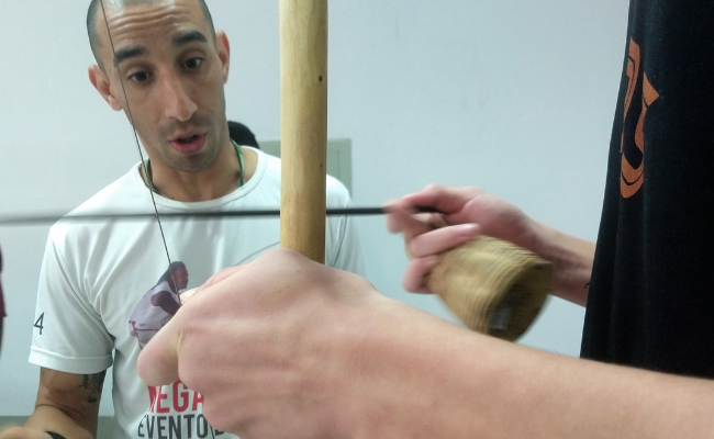Tatu, em pé, vestindo uma camisa branca, e um berimbau