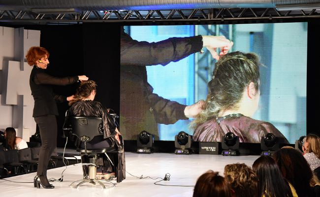 Em um palco aberto, com uma cadeira no meio, uma cabelereira mexe no cabelo de uma cliente. No fundo, um telão mostra estes movimentos no cabelo