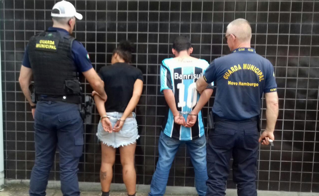Em pé, ao centro, uma mulher e um homem algemados com as mãos para trás. Dois Guardas, um de cada lado, fazem a segurança