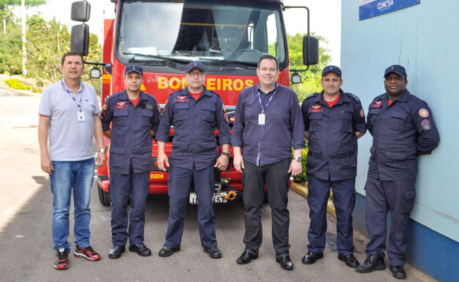 Seis homens em pé lado a lado, sendo cinco com o fardamento dos bombeiros. Um caminhão de bombeiros atrás do grupo