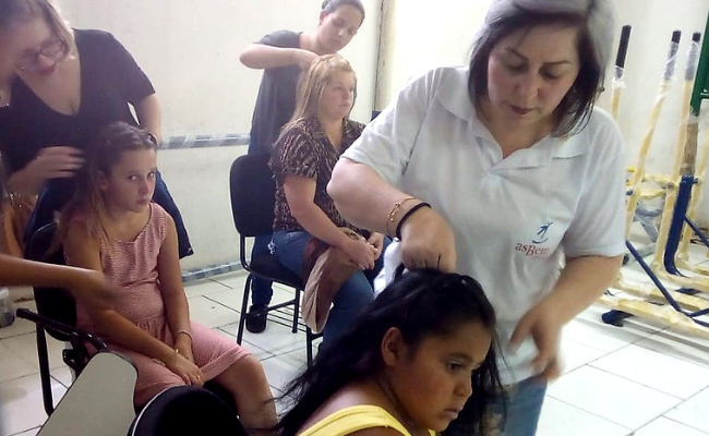 Uma menina sentada, com uma instrutora em pé mexendo com um escova o seu cabelo. Ao fundo, mais duas meninas sentadas també recebendo o tratamento no cabelo