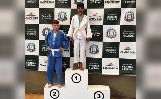 Em um pódio com três lugares, sendo o primeiro mais alto, um menino em pé de quimono branco está no primeiro lugar e um segundo menino de quimono azul ocupa o segundo lugar, que é à direita