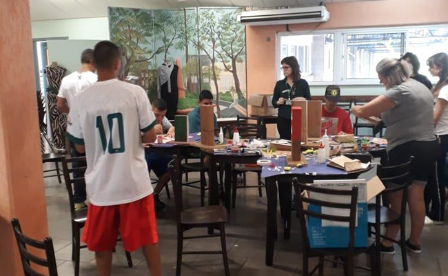 Em uma sala, pessoas em pé no entorno de uma mesa, que está com objetos em cima