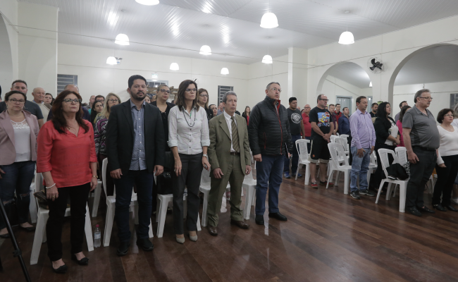 Em um salão, público de frente e em pé, com os secretários formando a primeira fila lado a lado