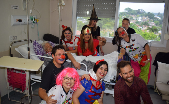 Pousados para a foto, grupo Caçadores de Sorrisos ao lado e a frente de uma cama hospitar, com uma senhora sentada