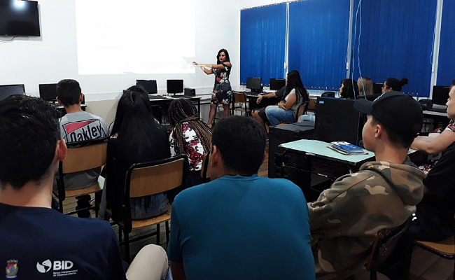 Em uma sala de aula, muher em pé ao fundo e alunos sentados a sua frente