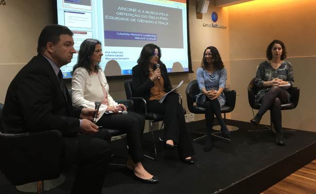 Em um palco, cinco pessoas sentadas em cadeiras organizadas lado a lado. A secretaria Paraskevi está sentada na segunda cadeira da esquerda para a direita. Um telão faz a projeção de uma apresentação atrás das cadeiras