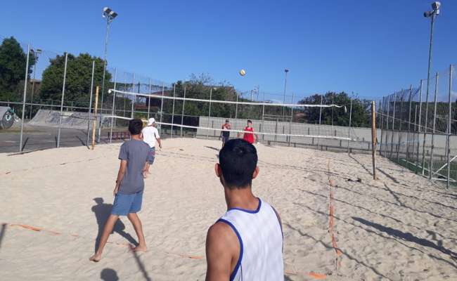 Quadra de areia, com um rede no meio, e dois jogadores homens de cada lado