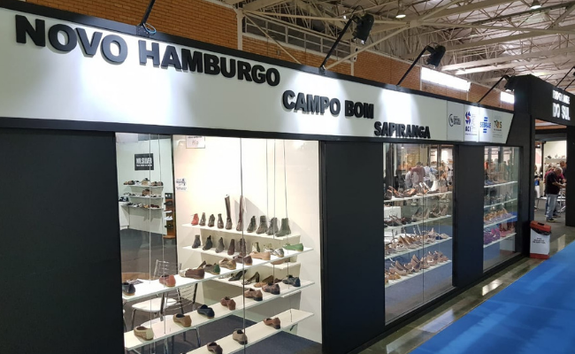 À esquedar, estande com pilares pretos e de vidro no centro, com calçados expostos. Acima, uma placa escrita Novo Hamburgo, Campo Bom e Sapiranga