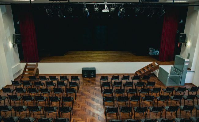 No fundo, o palco multiuso. Em frente ao palco, cadeiras posicionadas em filas